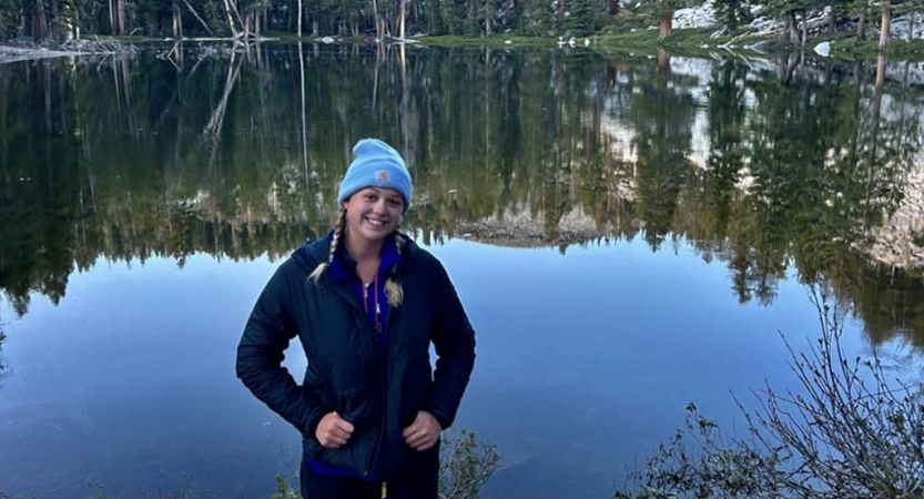 A person stands near a body of water, which is reflecting the trees surrounding it. 